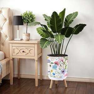 May include: A white cylindrical planter with a floral design, supported by three wooden legs. The planter features a watercolor pattern of pink, blue, and yellow flowers. A large green plant with broad leaves is inside the planter.