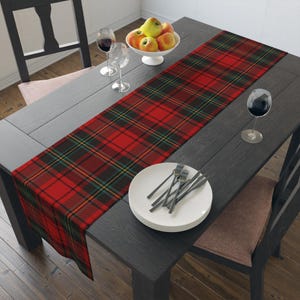May include: A red and black plaid table runner is displayed on a dark gray wooden table. The runner features a classic tartan pattern with green and yellow accents. A white bowl of apples, wine glasses, and a plate with silverware are also on the table.