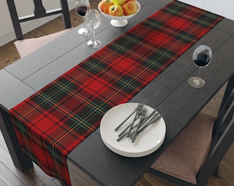 May include: A red and black plaid table runner is displayed on a dark gray wooden table. The runner features a classic tartan pattern with green and yellow accents. A white bowl of apples, wine glasses, and a plate with silverware are also on the table.