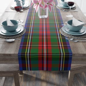 May include: A colorful plaid table runner in red, green, blue, yellow, and white. The runner is placed on a wooden table, with place settings and a vase of pink flowers. The plaid pattern is a classic design.