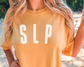 May include: A mustard yellow t-shirt with the text "SLP" printed in white on the front. The t-shirt is being worn by a person on a beach.