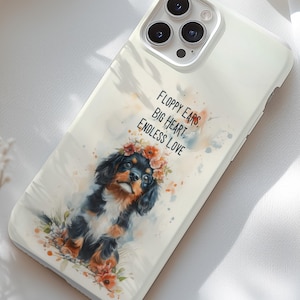 Może przedstawiać: Kremowe etui na telefon z akwarelową ilustracją Cavalier King Charles Spaniel w koronie z kwiatów. Na etui widnieje napis "Floppy Ears, Big Heart, Endless Love". Etui posiada wycięcie na aparat z trzema obiektywami.