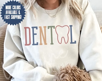 May include: A light gray sweatshirt with the word "DENTAL" in colorful letters and a tooth graphic. The sweatshirt is being worn by a person and a small dog is sitting on their lap. The text "COLOR SAND" is visible at the bottom of the image.