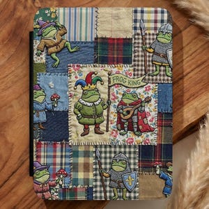 May include: A patchwork journal with frog illustrations in different costumes. The cover is made of various fabric patterns, including plaid and floral designs. The phrase "FROG KING" is visible on a banner held by a frog.
