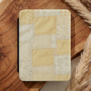 May include: A tablet case with a patchwork design in shades of yellow and white. The case features a variety of fabrics, including floral and gingham patterns. The case is set on a wooden chopping board with dried ornamental grass.