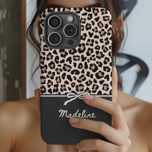 May include: A black phone case with a leopard print design and a white bow. The case has a black bottom section with the name "Madeline" written in white.