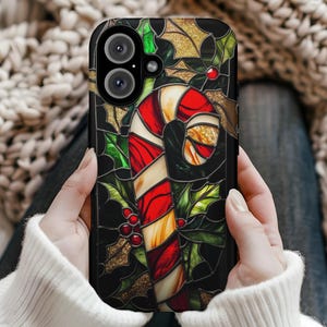 May include: A black phone case featuring a stained-glass design of a red and white candy cane, holly leaves, and berries. Gold glitter accents the design. The case has a dual-lens camera cutout.