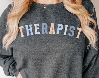 May include: A dark grey sweatshirt with the word "THERAPIST" printed in rainbow colours across the chest.