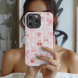 Op de afbeelding: Een roze en witte telefoonhoes met een ruitjespatroon en kersen- en strikillustraties.