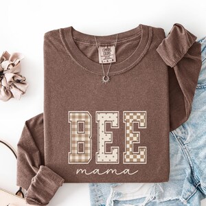 May include: A brown long-sleeve crewneck sweatshirt featuring the word BEE in large block letters with varied patterns including gingham, polka dots, and checkerboard, with the word mama in white cursive script underneath the block letters.
