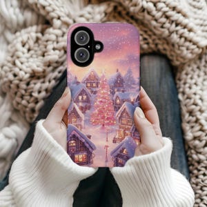 Puede incluir: Una funda de teléfono con una escena de un pueblo invernal con casas cubiertas de nieve, un árbol de Navidad rosa y un cielo estrellado. La funda tiene un recorte para una cámara dual. La funda la sostiene una persona que lleva un jersey blanco.
