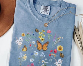 May include: A light blue t-shirt with a floral and butterfly print. The shirt has a crew neck and short sleeves. The text "COMFORT COLORS" is visible on the tag.