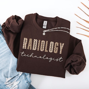 May include: A dark brown crewneck sweatshirt featuring the text RADIOLOGY in a beige and brown checkered pattern with small white flowers, and the word technologist written below in a white cursive script font, with a silver chain necklace.