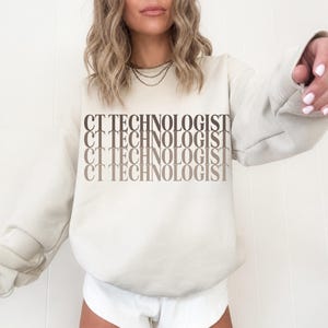 Op de afbeelding: Een beige sweater met ronde hals en de tekst CT TECHNOLOGIST vier keer onder elkaar gedrukt in een gestapeld kleurverloop van donkerbruin naar lichtbeige. Het kledingstuk met lange mouwen heeft een geribbelde kraag, boorden en zoom.