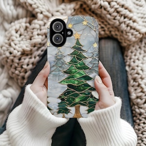 Puede incluir: Una funda de teléfono con un diseño de árbol de Navidad estilo vidriera. La funda tiene un árbol verde con detalles dorados, sobre un fondo gris nevado con estrellas doradas. La funda la sostiene una persona que lleva un jersey blanco.