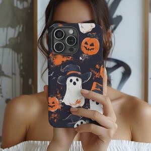 以下が含まれることがあります： ハロウィーンをテーマにしたデザインの黒い携帯電話ケース。オレンジ色のカボチャ、魔女の帽子をかぶった白い幽霊、オレンジ色のアクセントが付いた暗い背景が特徴です。ケースは光沢のある仕上がりです。