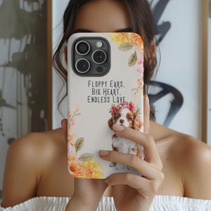 May include: A white phone case with a floral design and a picture of a Cavalier King Charles Spaniel puppy. The text on the case reads "Floppy Ears, Big Heart, Endless Love."