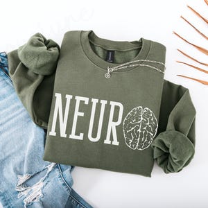 Può includere: Una felpa verde con la scritta "NEURO" e un'immagine di un cervello stampata sul davanti.