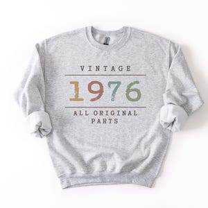 May include: A light grey crewneck sweatshirt featuring the text VINTAGE 1976 ALL ORIGINAL PARTS in dark brown lettering. The numbers 1976 are displayed in a distressed, multi-coloured font with shades of gold, rust, teal, and sage green.