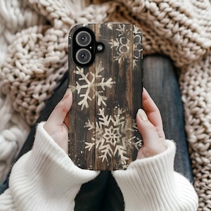 May include: A smartphone case featuring a rustic wood grain design with white snowflake patterns. The case has a dark brown background with light brown snowflakes. The phone case is held by a person wearing a white sweater.