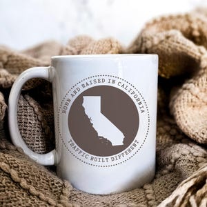 May include: A white ceramic mug featuring a circular graphic with a brown background and a white silhouette of the state of California. The text surrounding the circle reads BORN AND RAISED IN CALIFORNIA and TRAFFIC BUILT DIFFERENT.