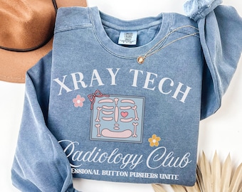 Puede incluir: Una sudadera azul claro con el texto "X-Ray Tech Radiology Club Professional Button Pushers Unite" y una imagen de dibujos animados de una radiografía de un esqueleto con un corazón.