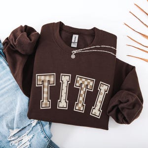 May include: A dark brown crewneck sweatshirt featuring the word TITI in large varsity-style letters. Each letter has a unique beige and brown pattern, including gingham, polka dots, checkerboard, and floral prints, with a white outline.