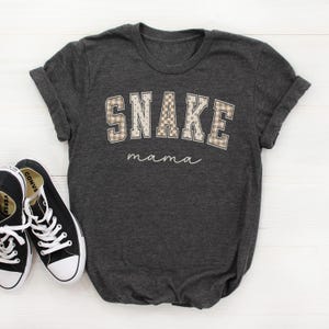 May include: A dark charcoal gray short-sleeve t-shirt featuring the word SNAKE in large, block-style letters with a beige and white checkered pattern, followed by the word mama in a white cursive script font, paired with black and white sneakers.