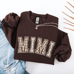 May include: A dark brown crewneck sweatshirt featuring the word MIMI in large block letters across the chest. Each letter has a unique pattern including beige and brown gingham, polka dots, checkerboard, and small floral prints with white outlines.