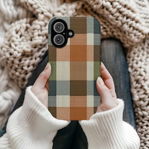 Pode incluir: Capa de smartphone com um padrão xadrez em tons de castanho, laranja, verde e creme. A capa tem um recorte para uma câmara de lente dupla. Uma pessoa com um suéter branco segura a capa.