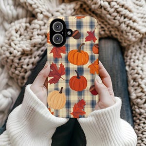 May include: A phone case featuring a fall-themed design. The case has a plaid background with orange pumpkins, red and orange leaves, and acorns. The case is held by a person wearing a white sweater.