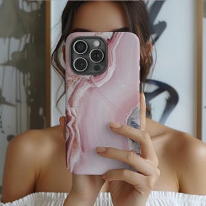 May include: A pink and white marble phone case with a camera lens cutout. The case has a swirling pattern reminiscent of agate.