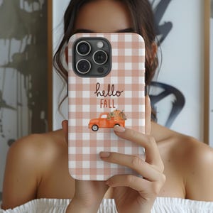May include: A phone case with a fall theme. The case has a peach and white gingham pattern. The words "hello FALL" are written in a script font. An orange truck carries pumpkins.