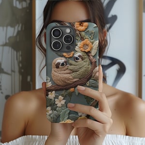 May include: A phone case with a floral design featuring two sloths embroidered on a branch. The case is light blue with green leaves and white flowers.
