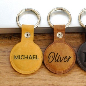 May include: Four leather keychains with metal rings. The keychains are brown and black and have different engravings. One keychain says "MICHAEL", another says "Oliver", another says "D|S", and the last one has a deer antler design with the letter "R" in the center.
