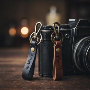 May include: Two leather keychains with metal clasps. One is black with "McAllister" engraved, the other is brown with "Reynolds" engraved. They are on a wooden surface with a vintage camera in the background.
