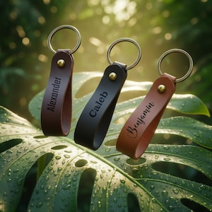 May include: Three personalized leather keychains in brown, black, and tan, each with a gold-colored ring and rivet. The keychains are engraved with the names "Alexander", "Caleb", and "Benjamin". The keychains are displayed against a backdrop of green leaves.