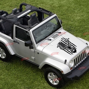 Op de afbeelding: Een zilveren Jeep Wrangler met een zwart-witte sticker van een hertenkop op de motorkap. De sticker heeft een Amerikaans vlagontwerp.