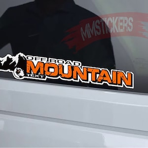 Può includere: Sticker decalco in testo Off-road Mountain in arancione e nero con una grafica di catena montuosa. Lo sticker è su un finestrino di un'auto.