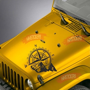 May include: A black and white compass with a mountain range design on a yellow Jeep hood. The compass has the cardinal directions N, S, E, and W, and the numbers 15, 30, 45, 60, 75, 105, 285, 300, 315, 330, and 345.