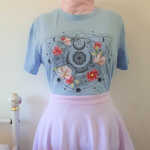 May include: A light blue t-shirt featuring a graphic print of a geometric design with a crescent moon, flowers, and circles. The t-shirt is worn with a lavender skirt with a white lace trim.