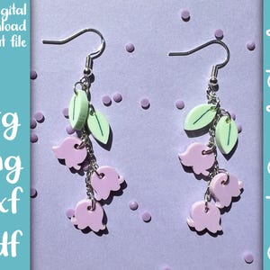 Op de afbeelding: Een paar oorbellen met een cluster van drie lichtpaarse lelietjes-van-dalen bloemen met groene bladeren die aan een zilveren ketting hangen. De oorbellen zijn op een lichtpaarse achtergrond.