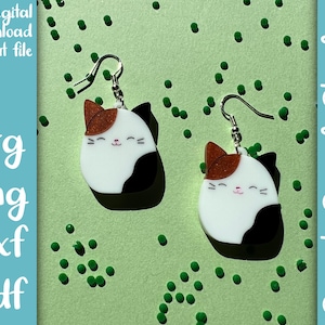 May include: A pair of cat-shaped earrings with a white, black and brown colour scheme. The earrings are on a green background with small green dots.