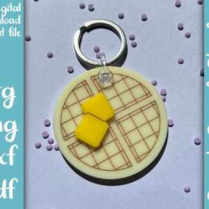 May include: A white waffle keychain with a yellow butter pat on top. The keychain is attached to a silver key ring.