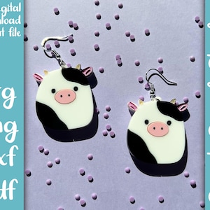 May include: A pair of black and white cow-shaped earrings with pink noses and gold accents. The earrings are on a purple background with small purple dots.