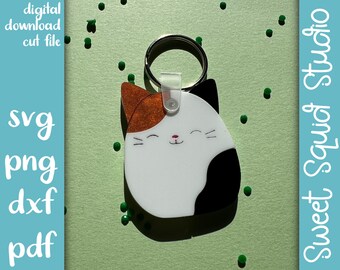 May include: A white, black, and orange cat keychain with a smiling face. The keychain is attached to a silver key ring.