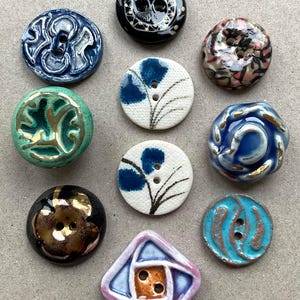 Puede incluir: Una colección de diez botones de cerámica de diversas formas, colores y patrones. El conjunto incluye botones redondos y cuadrados con acabados esmaltados en azul, verde, negro, blanco y rosa, con detalles decorativos en dorado, marrón y negro.