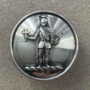 Puede incluir: Un botón circular de tono plateado con un diseño en relieve de una figura con armadura histórica. La figura lleva un casco, sostiene un bastón rematado con una cruz en la mano derecha y se apoya sobre una base trenzada con una espada al lado.