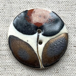 May include: A round ceramic button featuring a glossy off-white base with a hand-painted abstract design in shades of deep black, metallic bronze, and copper. The button has two central holes and a smooth, reflective finish across the surface.