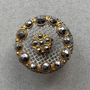 May include: A circular button featuring a silver-toned metal mesh base with a gold-toned metal border and center floral motif. The design is embellished with multiple faceted dark gray stones set along the outer edge and the central flower.